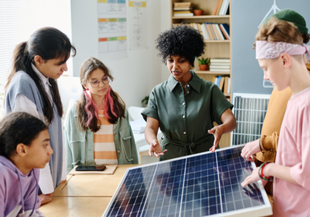 Students gather around a solar panel in a classroom, examining it and discussing its features.