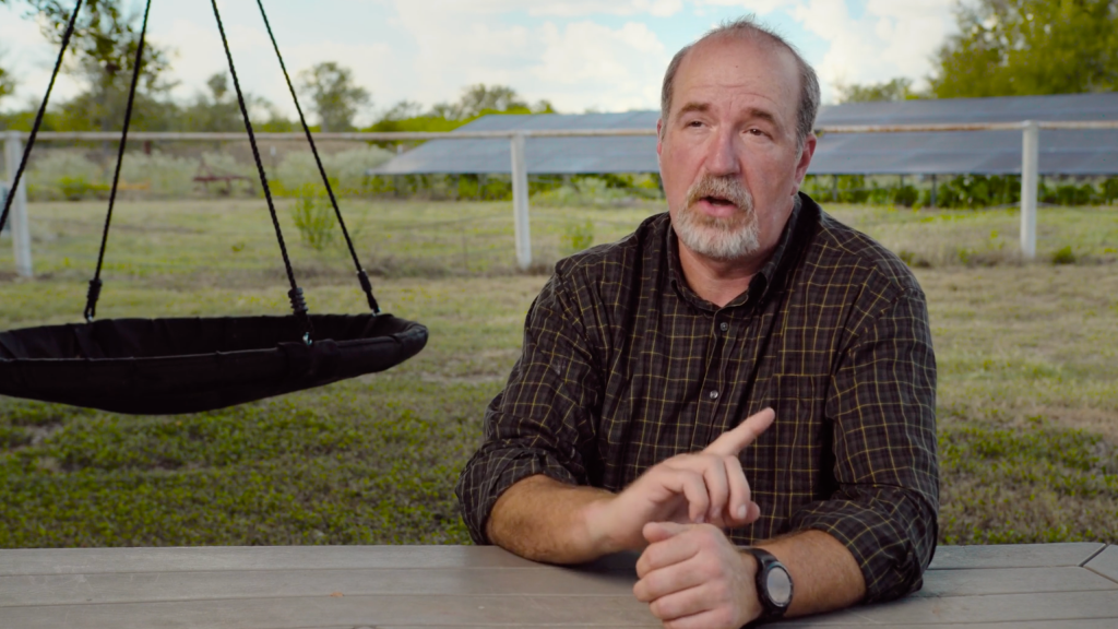 A man in a plaid shirt sits at a wooden table, gesturing with his hand, with a black swing hanging in the background and a fence and buildings beyond.