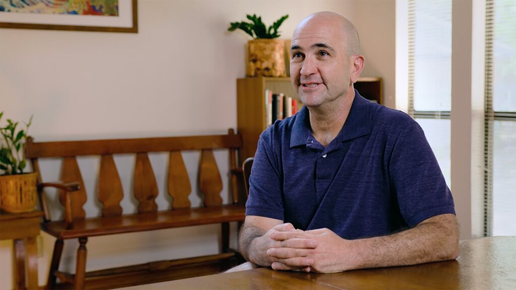 A man in a dark blue polo shirt sits at a wooden table, hands clasped, with a wooden bench and a framed picture on the wall behind him.