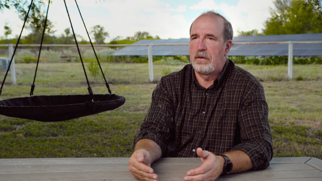 A man in a plaid shirt sits at a wooden table, hands clasped, with a black hanging chair nearby and a fence and building in the background.