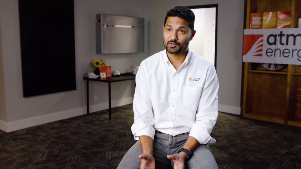 A man in a white shirt and gray pants sits on a carpeted floor, hands near his knees, with a black wall featuring a white panel and a red and white "atm energy" sign behind him.