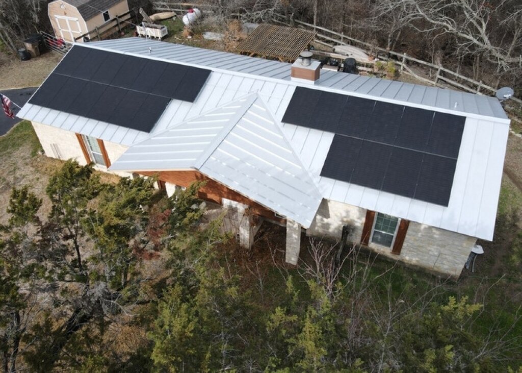 A white house with black solar panels and a wooden deck is surrounded by a natural forest backdrop.