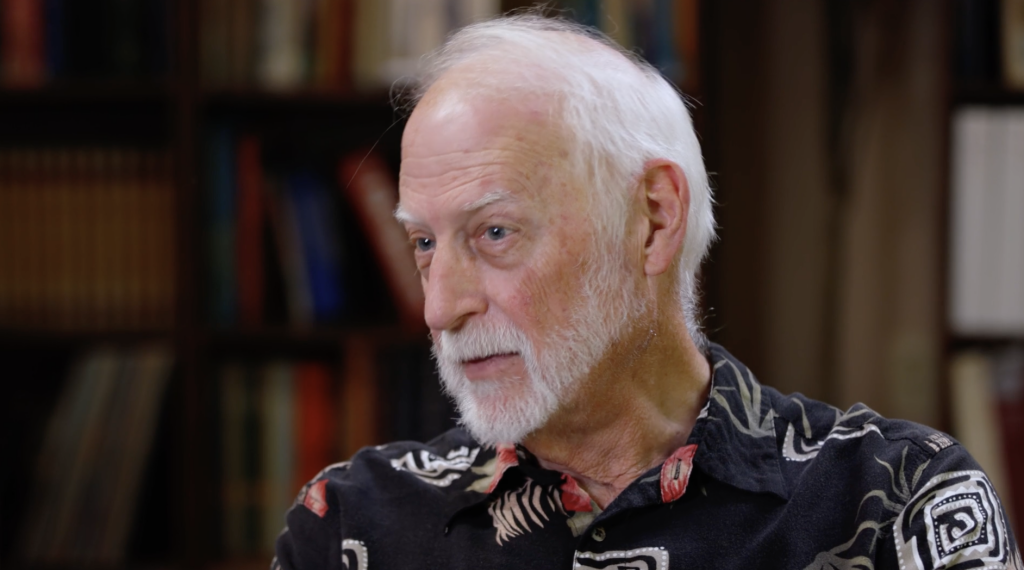 An older man with white hair and beard, wearing a black shirt with a white pattern, sits in front of a bookshelf, looking off to the side.