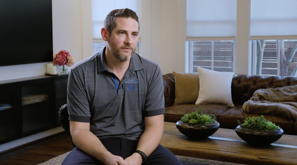 A man in a gray polo shirt and dark pants sits on a wooden coffee table in a living room with a brown leather couch, two potted plants, a black TV stand, and a large window with white blinds.