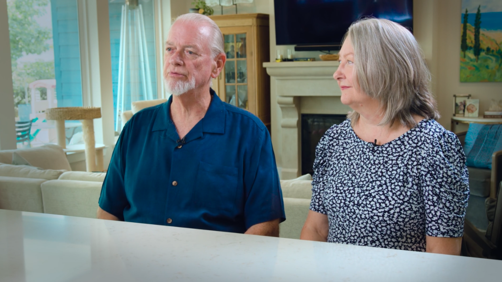 An older man in a blue shirt and a woman in a blue and white patterned dress sit together on a white table, with a fireplace, window, cat tree, and painting visible in the background.