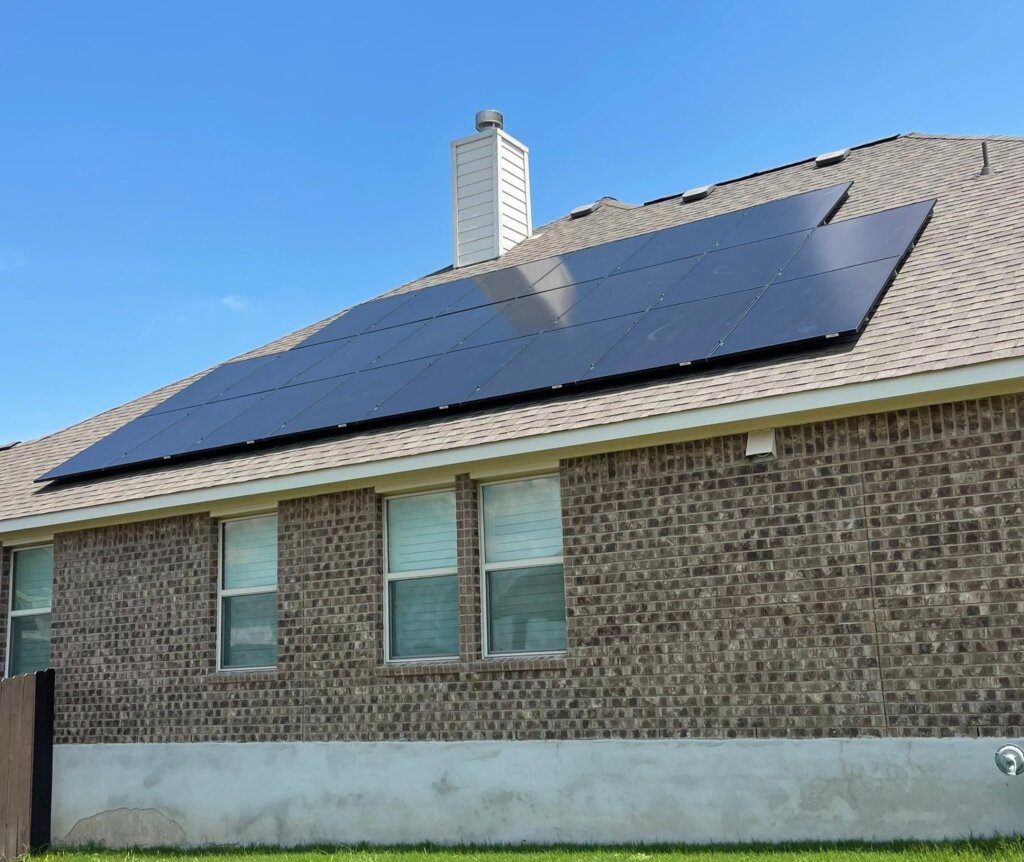 A house with gray roof and brick exterior features two solar panels and a white chimney, with a blue sky and grassy area in the background.