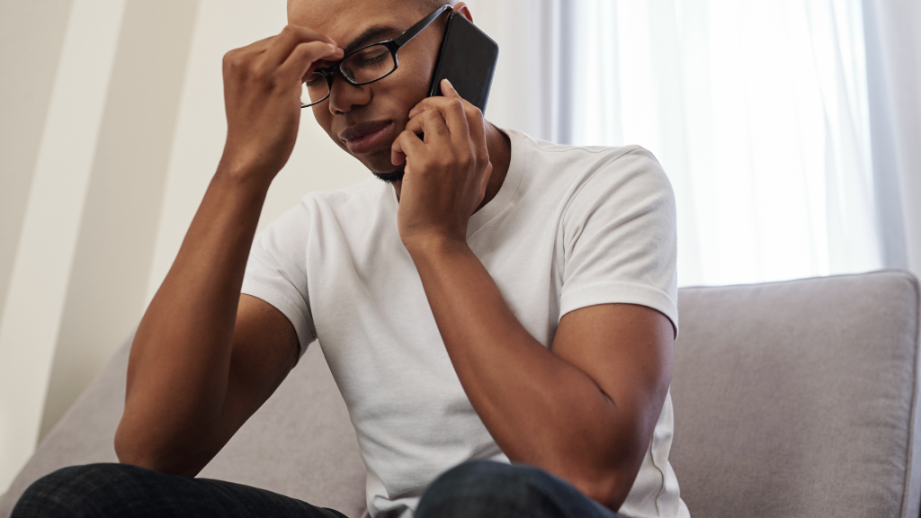 A man wearing glasses and a white t-shirt sits on a gray couch, talking on a black cell phone.