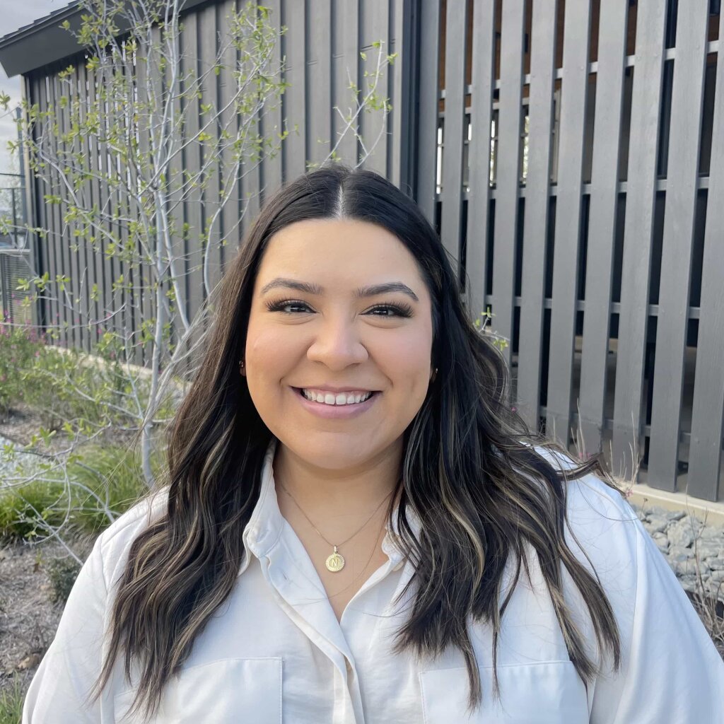 A young woman with long, dark hair and a white shirt smiles warmly, wearing a gold necklace in front of a gray wooden fence with a tree nearby.