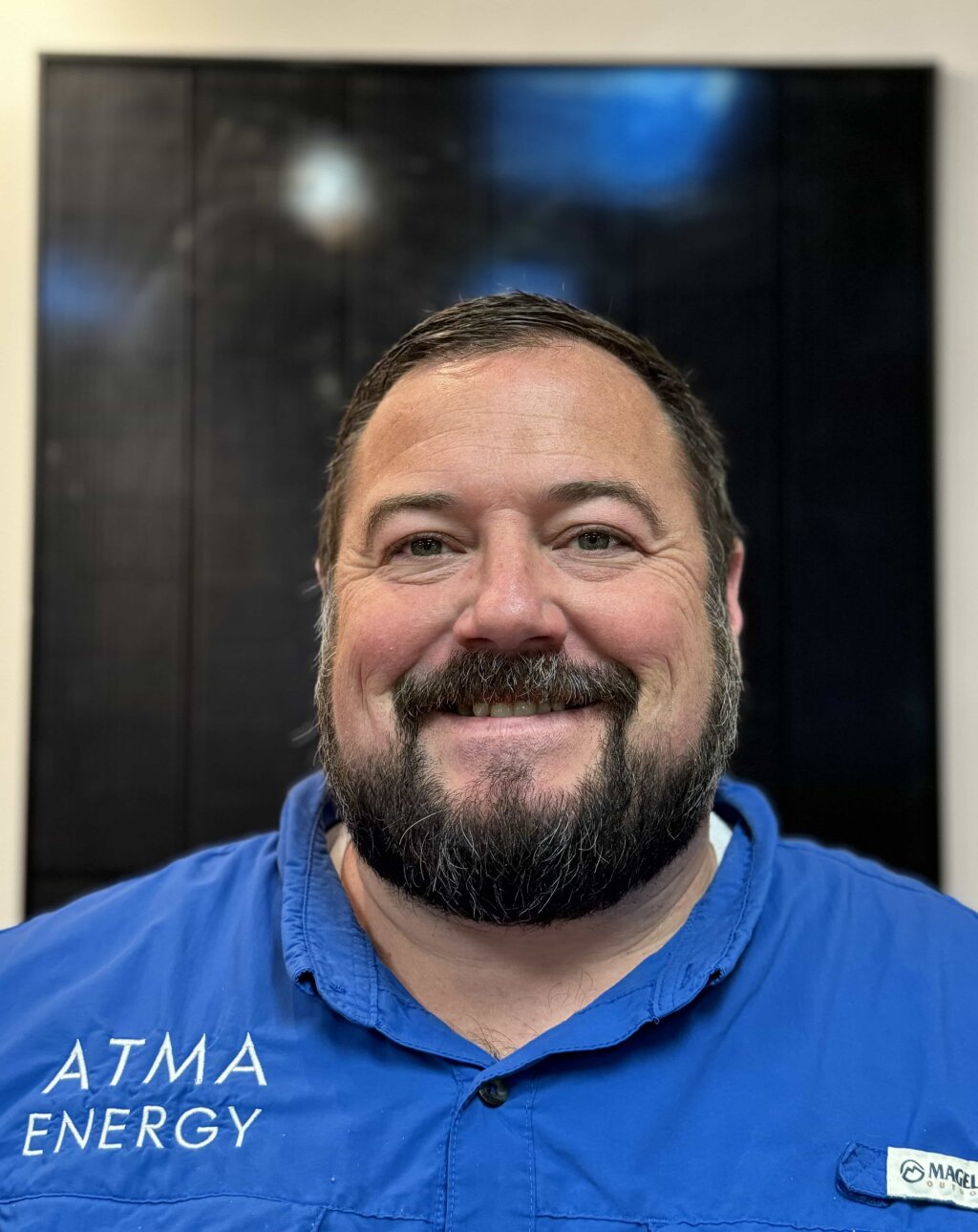 A man in a blue shirt, wearing a name tag with "ATMA ENERGY", smiles in front of a black screen displaying a white message.