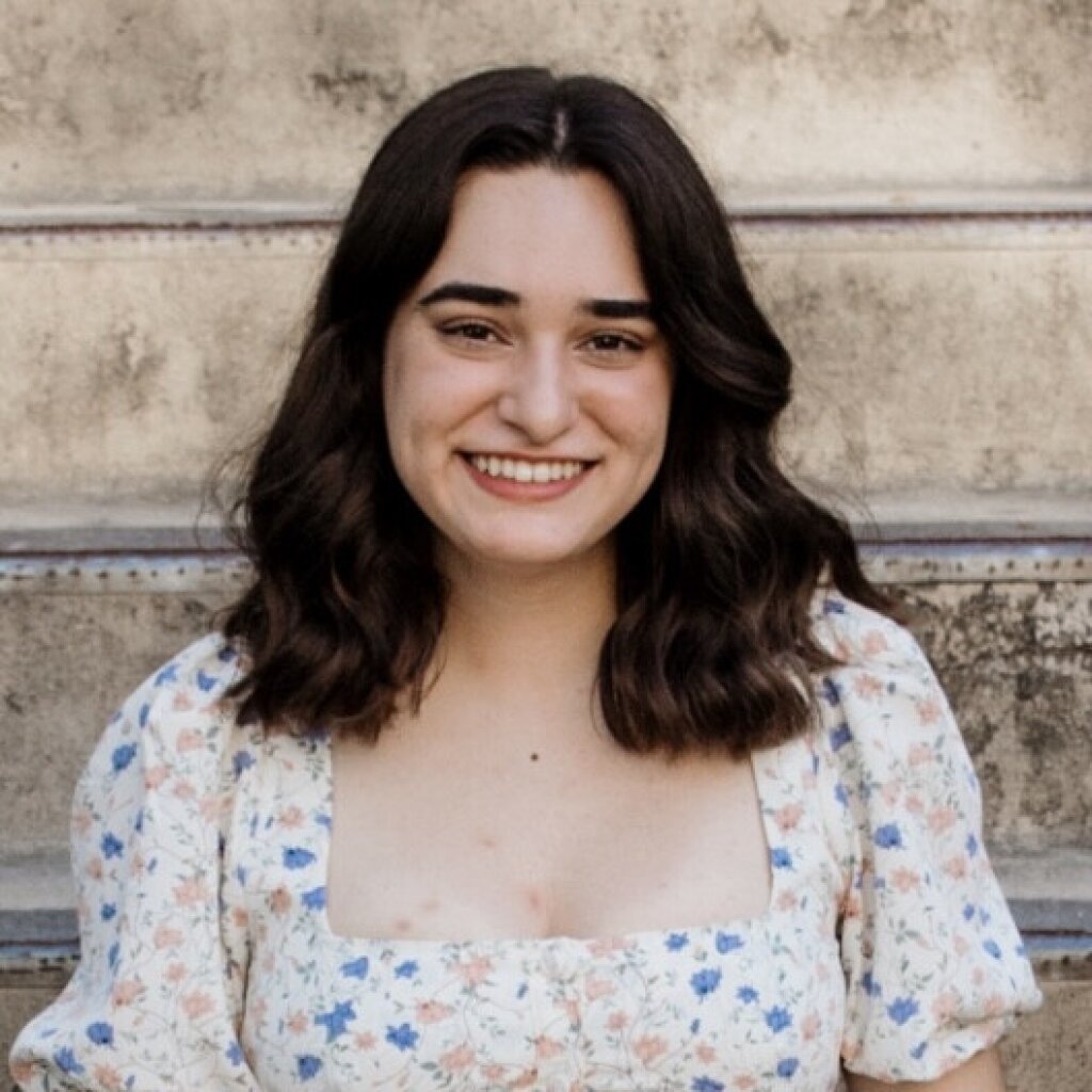 A young woman with dark hair and a radiant smile wears a white dress with blue and pink flowers, positioned against a beige wall with concrete columns.