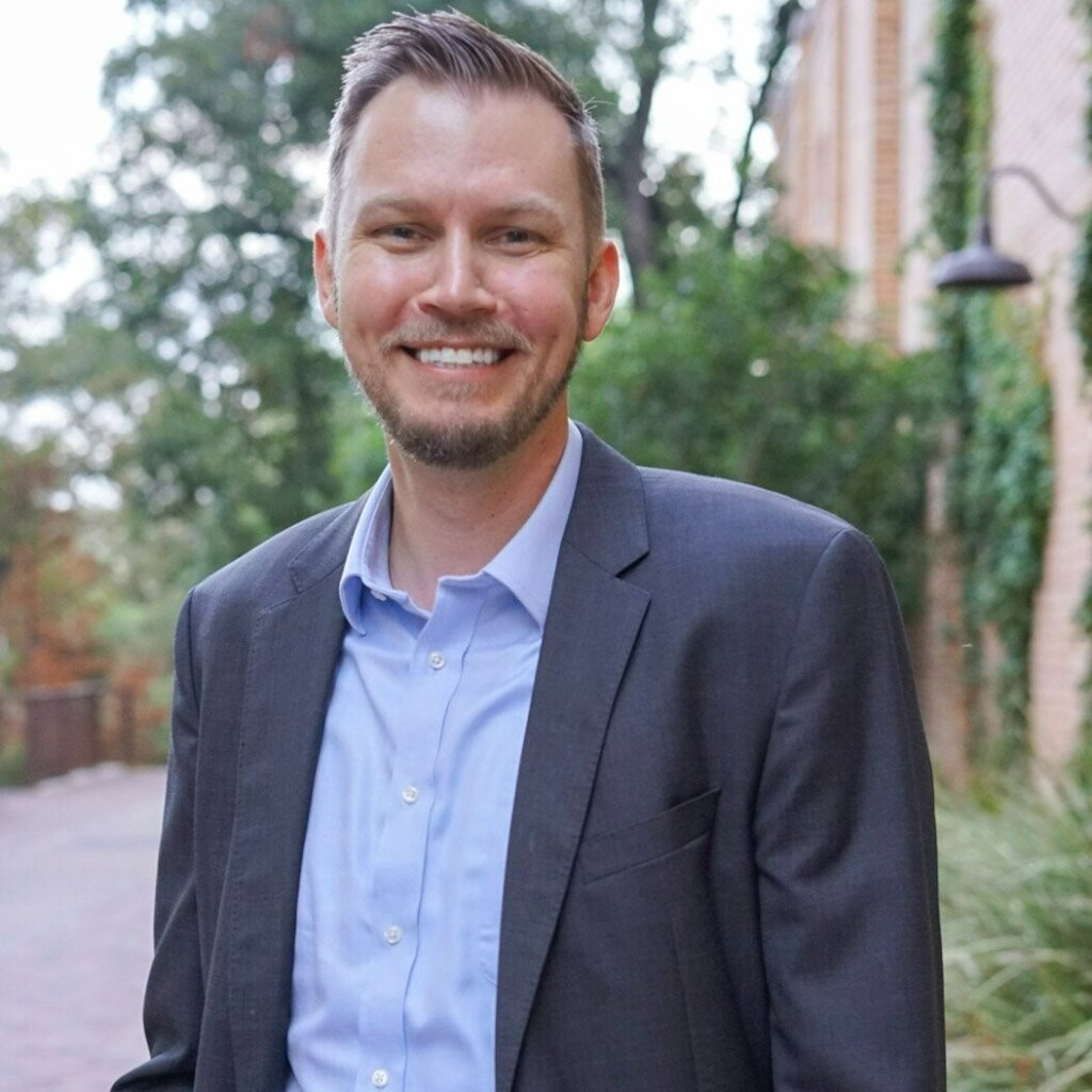 A man in a dark suit and light blue shirt smiles in front of a brick building on a tree-lined street.