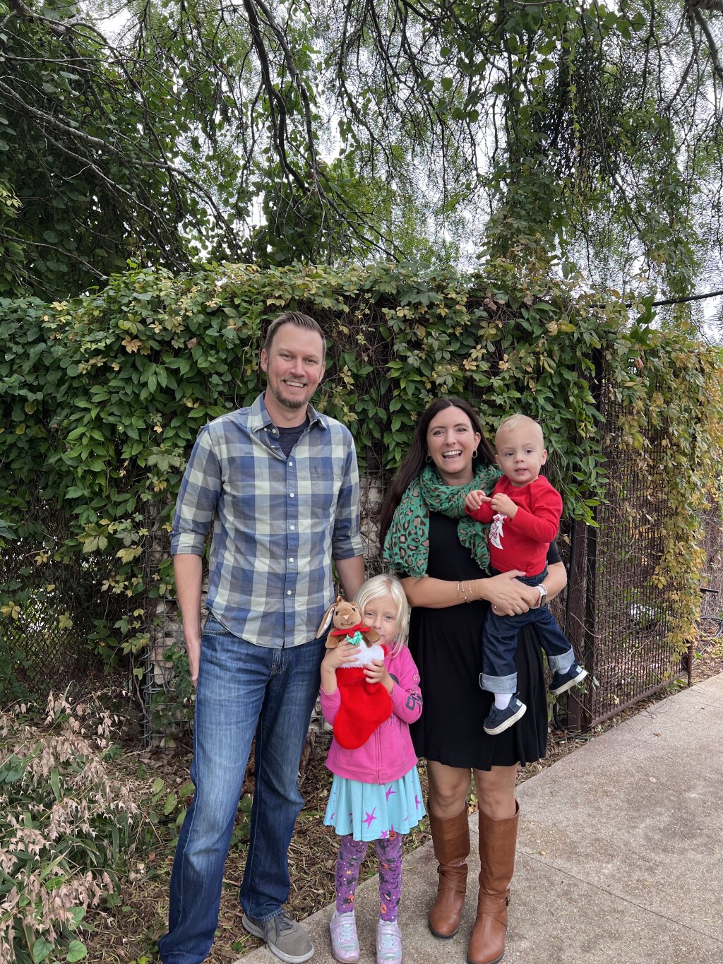 A family of four, dressed casually and posing for a photo, stands in front of a chain link fence with a tree and bushes behind them.