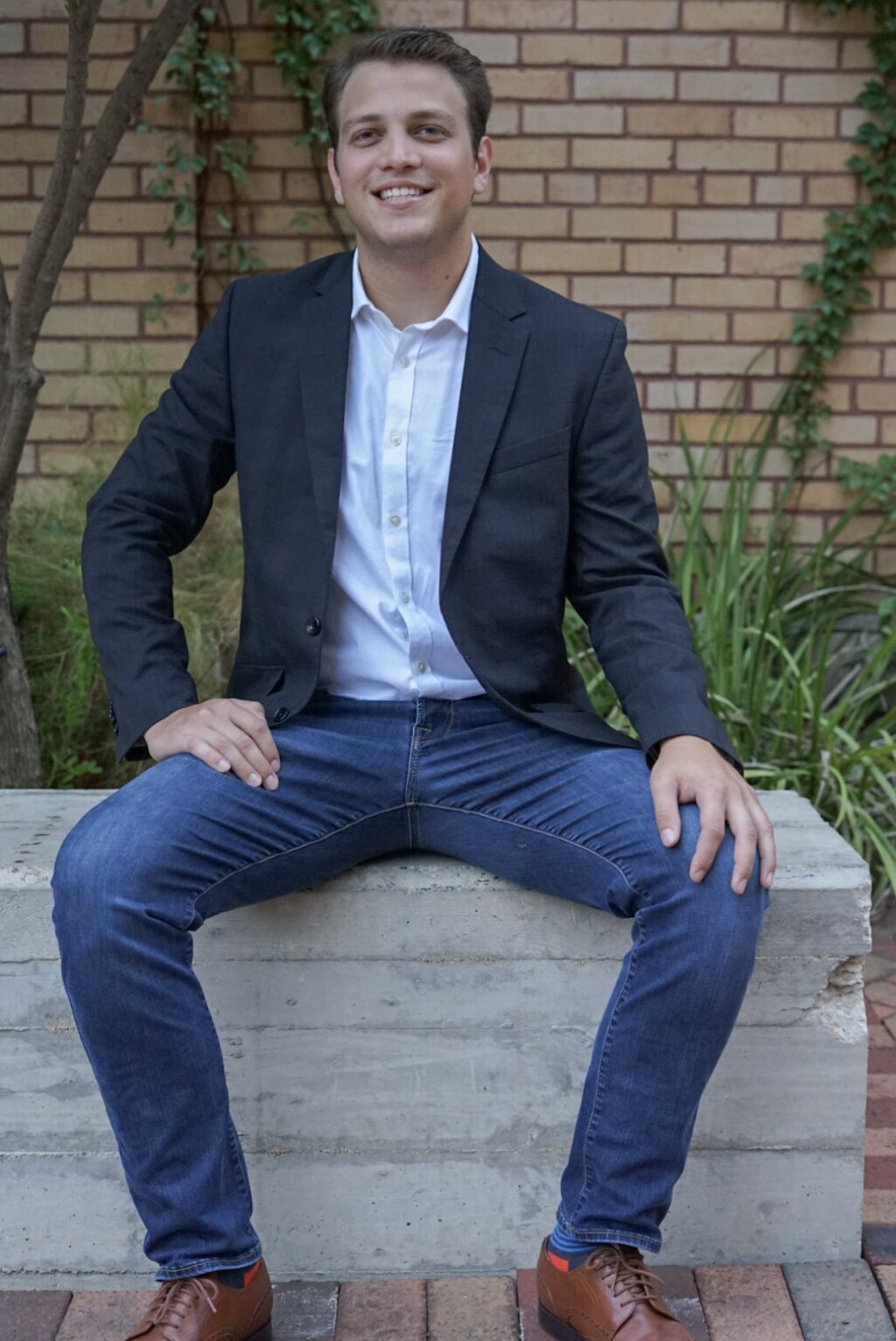 A young man in a navy blazer, white shirt, and blue jeans smiles, seated on a concrete bench with a brick pathway and greenery in the background.