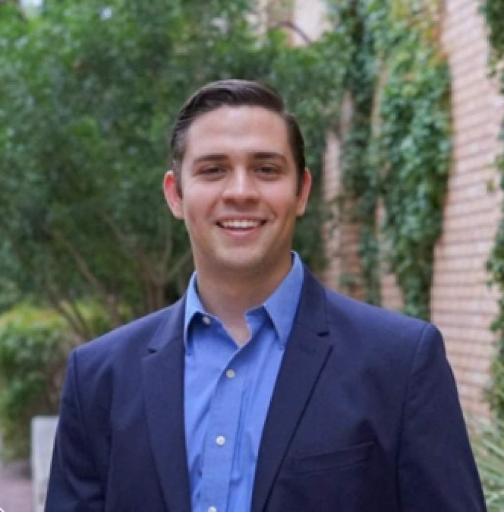 A young man in a blue suit jacket and shirt smiles warmly in front of a brick wall with greenery.