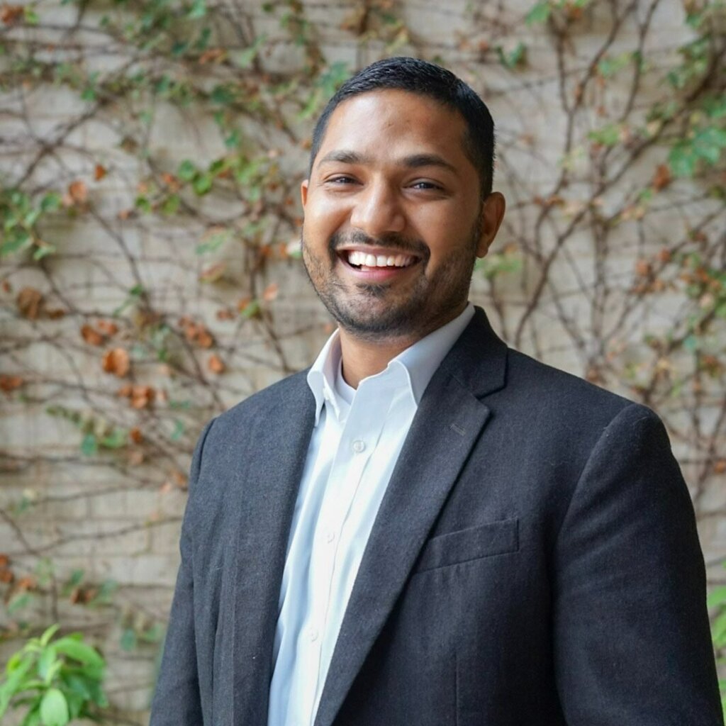 A man in a dark gray suit, white shirt, and dark tie smiles in front of a brick wall with green ivy, creating a striking contrast.