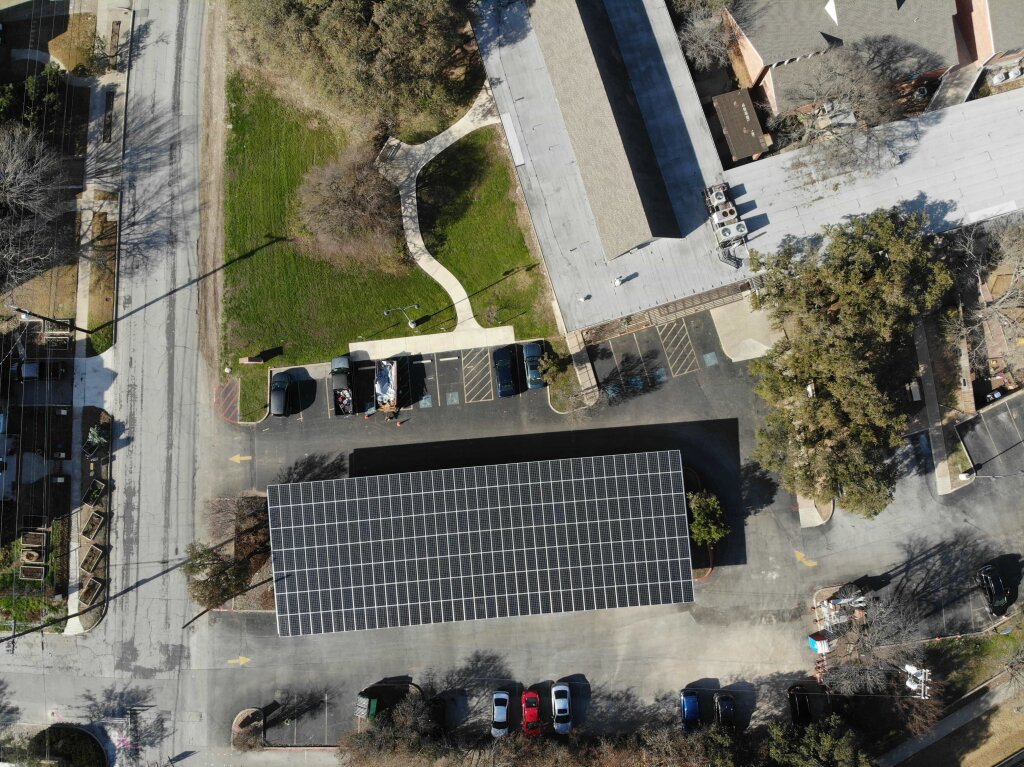 An aerial view of a residential area reveals a solar-paneled building, empty parking lot, and a mix of houses, trees, and greenery.