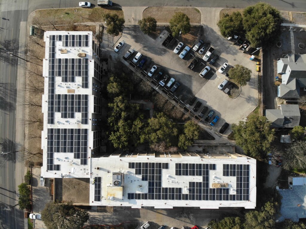 An aerial view of a cityscape showcases two buildings with solar panels, a parking lot, and a prominent crosswalk, framed by trees.