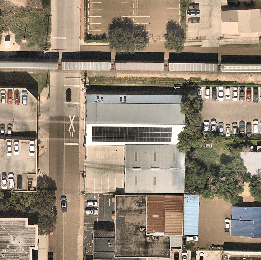 An aerial view of a city street showcases parked cars, a large building, and trees, with patches of green grass and trees interspersed.