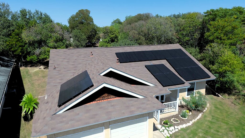 A house with brown shingled roof and black solar panels is situated on a lush green lawn, surrounded by trees and a white picket fence.