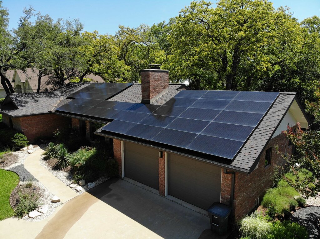 A modern brick house with gray shingled roof and black solar panels is situated on a concrete driveway, surrounded by lush greenery and a small garden.