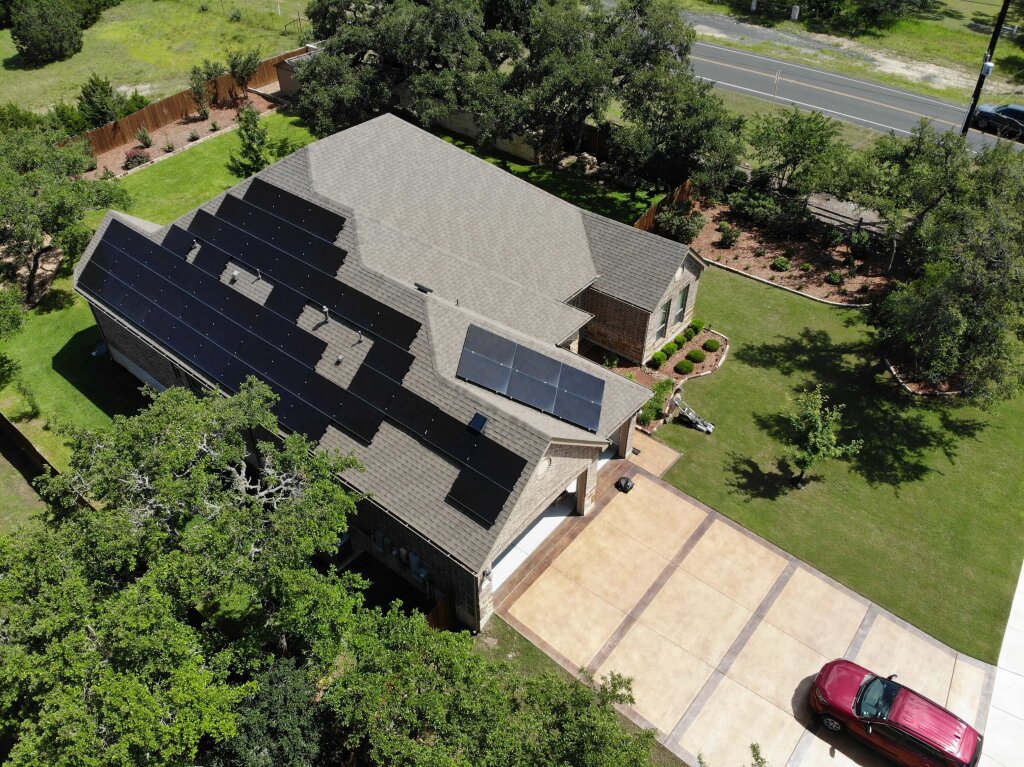 A gray house with solar panels and a brick driveway is surrounded by a lush green lawn, a red car, and a solitary tree.