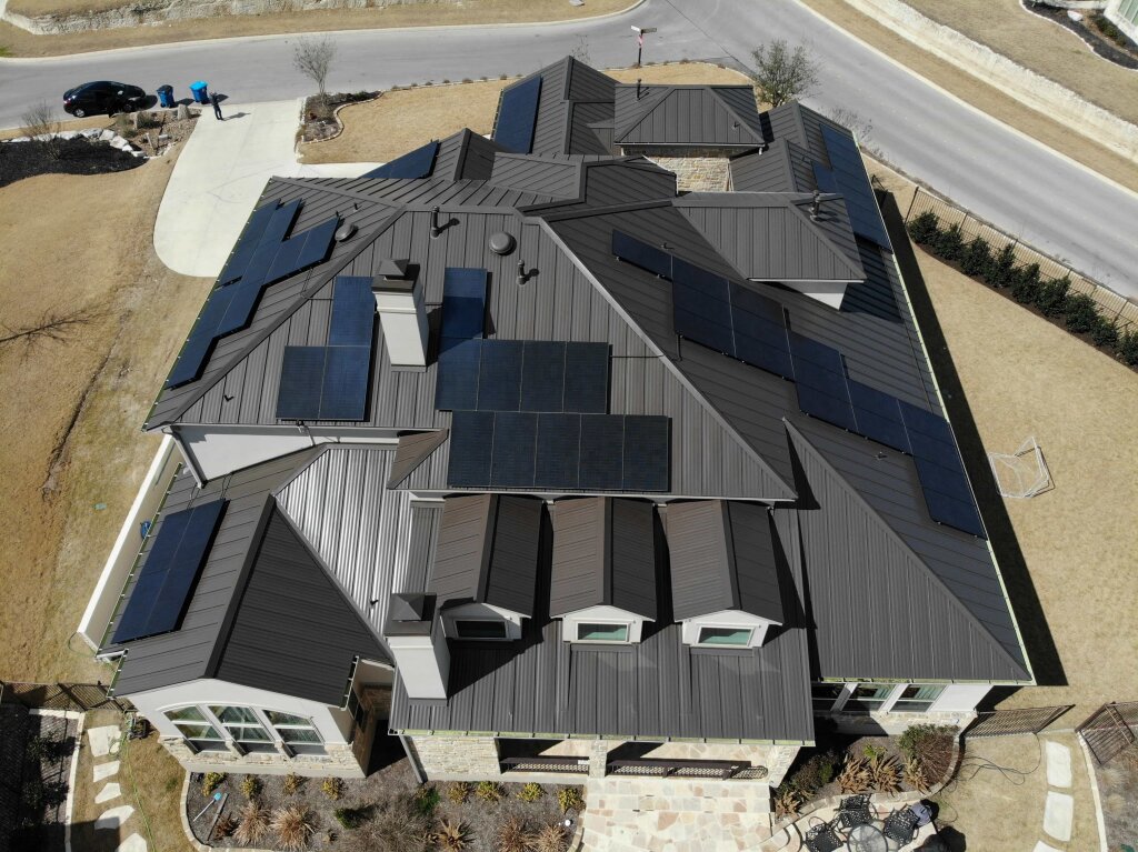 A modern two-story house with numerous solar panels covers a metal roof, situated on a suburban street with a sidewalk and lawn.
