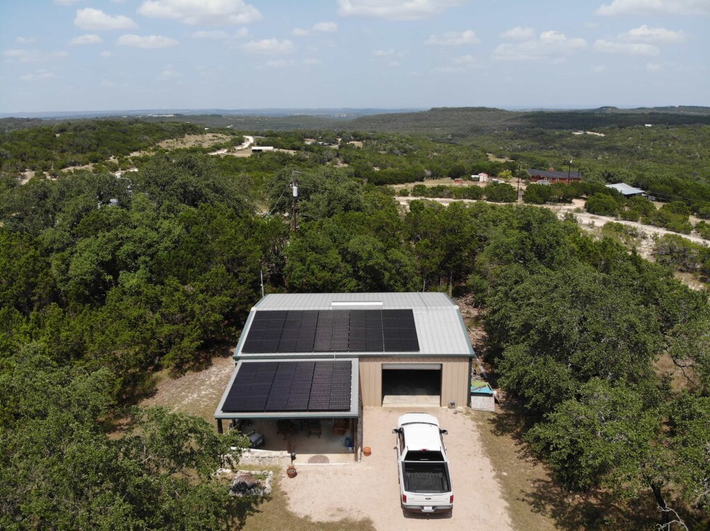 A beige house with solar panels and a white truck parked in the driveway is situated in a rural landscape, surrounded by trees and rolling hills.