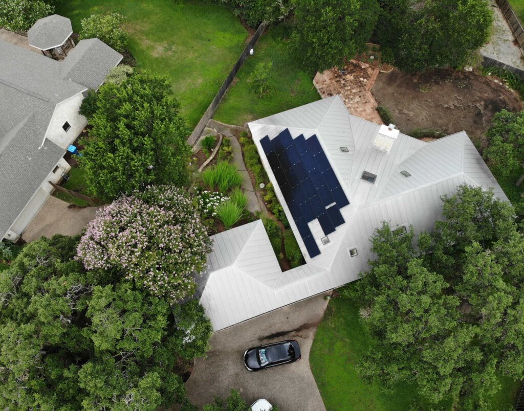 A white house with solar panels and a gray roof is surrounded by a lush green lawn, a garden, and a black car in the driveway.